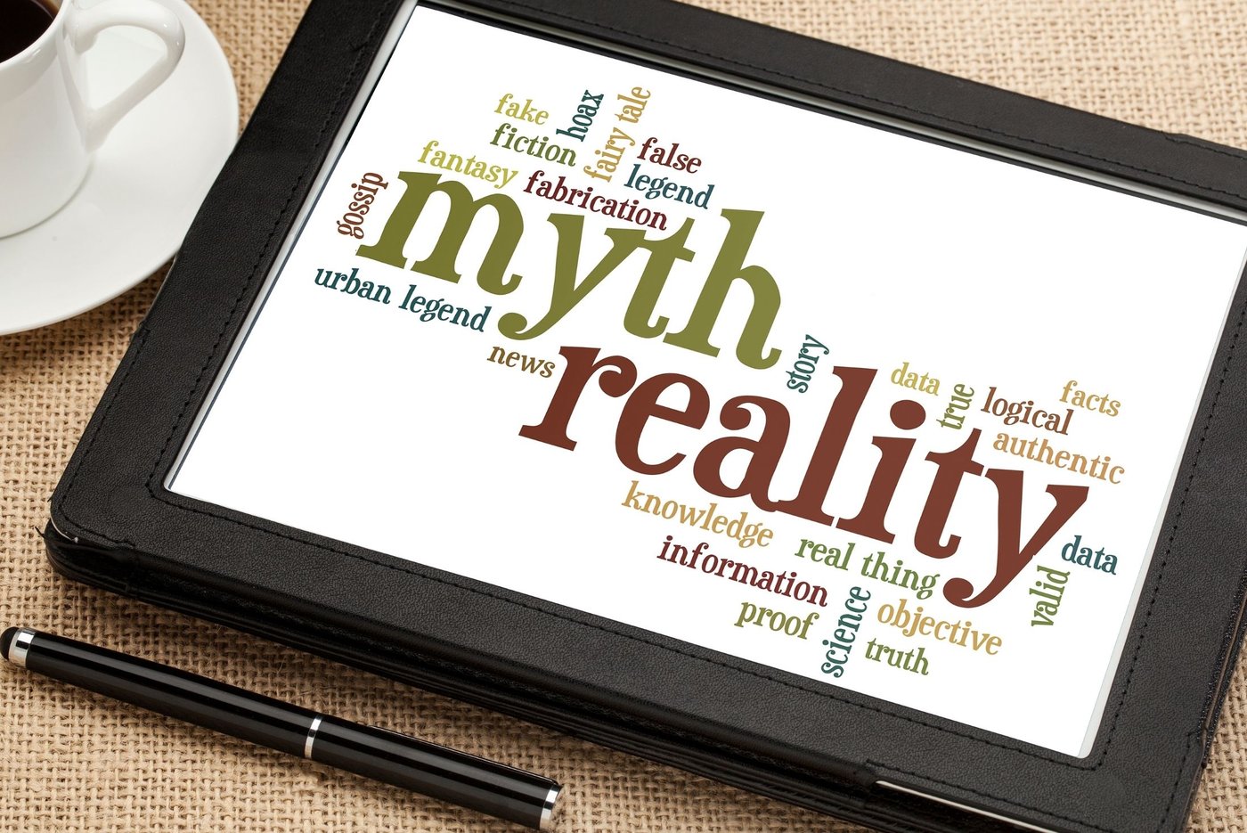 Person mit Kaffee und Tablet mit einer Tagcloud rund um das Thema Mythen und Realität – Mythen beim Start ins Coaching-Business